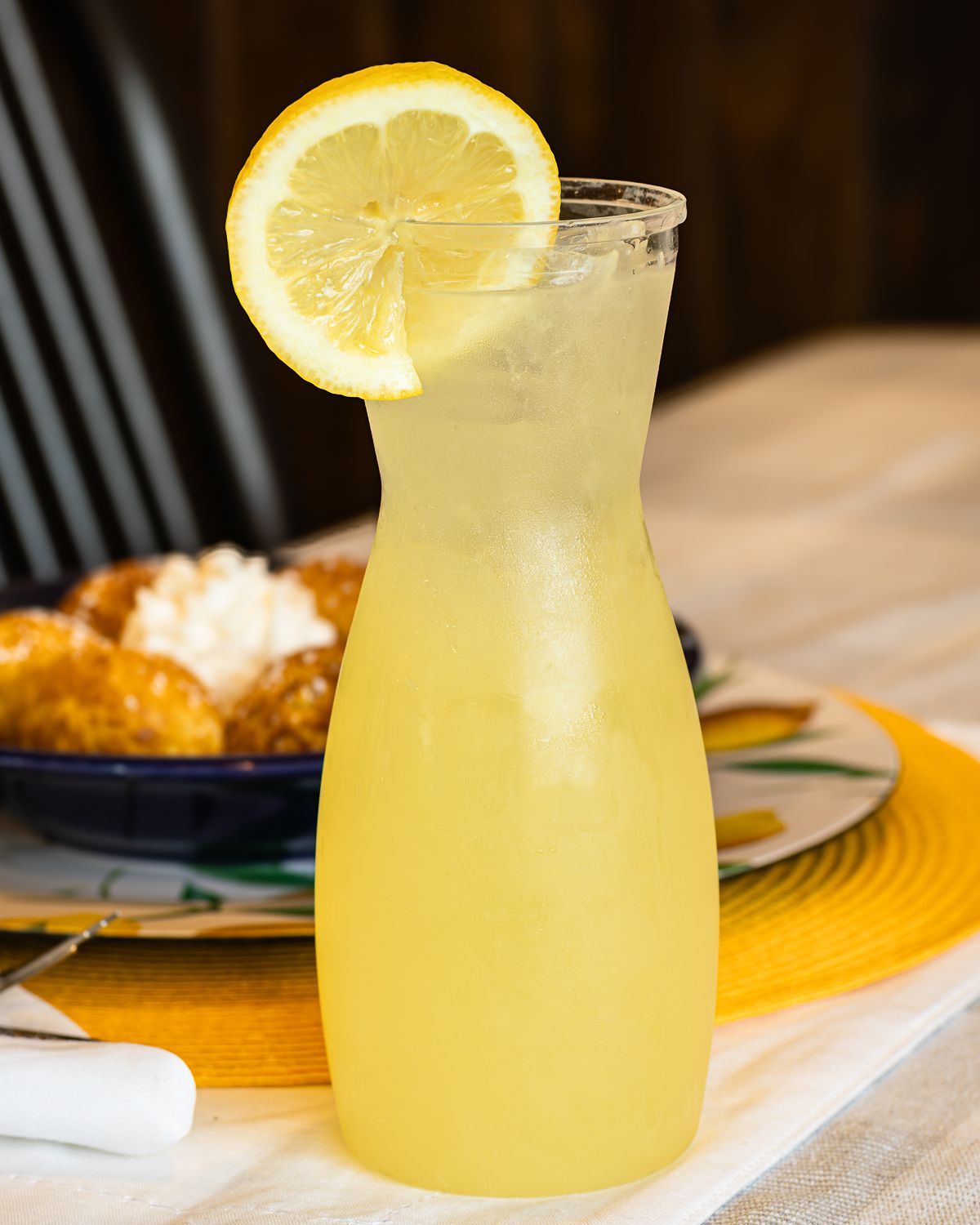 Pitcher of yellow lemonade with a lemon slice garnish, sitting on a table with pastries and a yellow placemat.