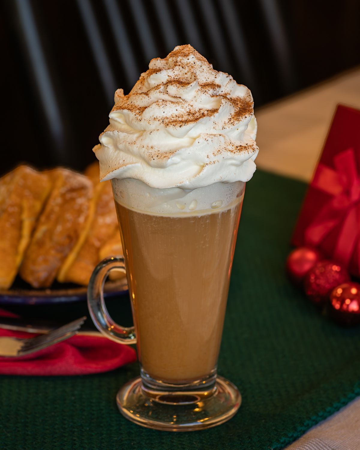 Latte with whipped cream and cinnamon, served in a tall glass; bread and a gift are in the background.