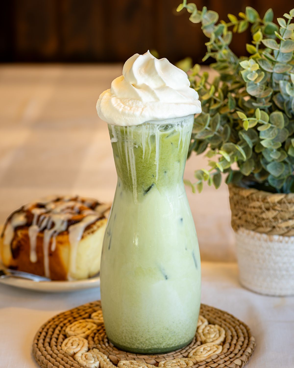 Iced matcha latte topped with whipped cream in a decorative glass, with a cinnamon roll and plant in the background.