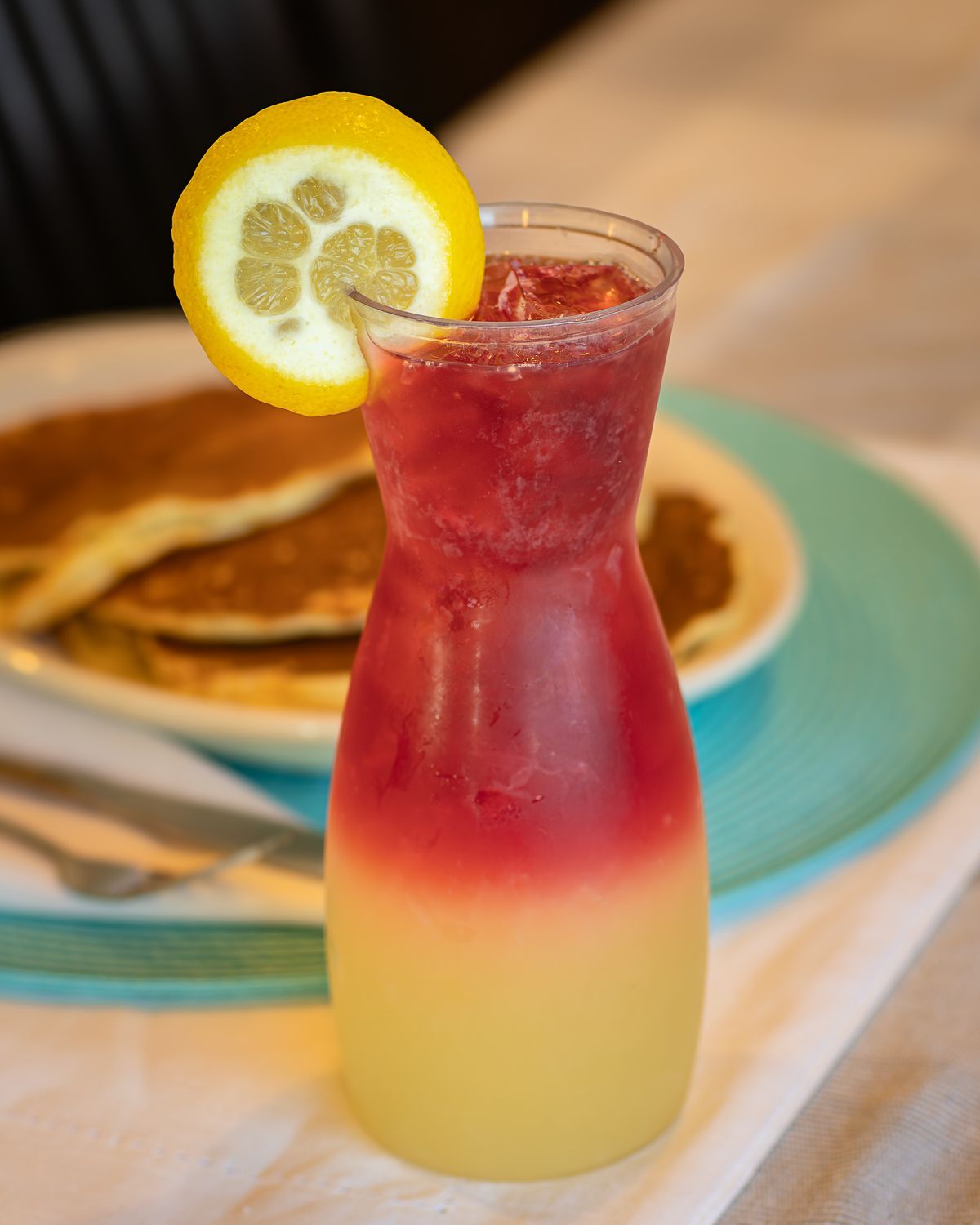 Glass of layered drink with a lemon slice garnish, in front of a plate of pancakes.