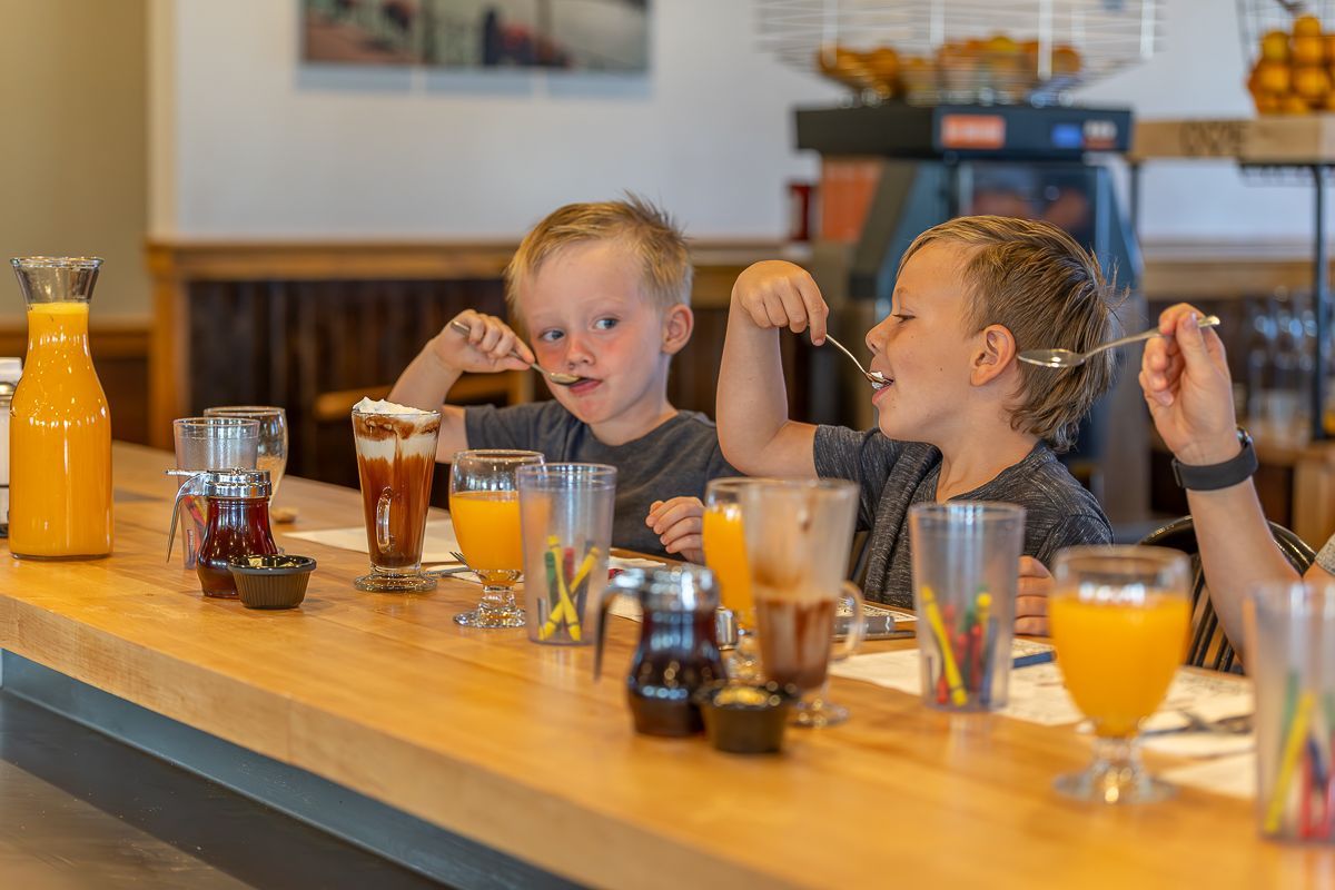 Two children eating from spoons at a diner counter. Orange juice, drinks, and syrups are on the counter.