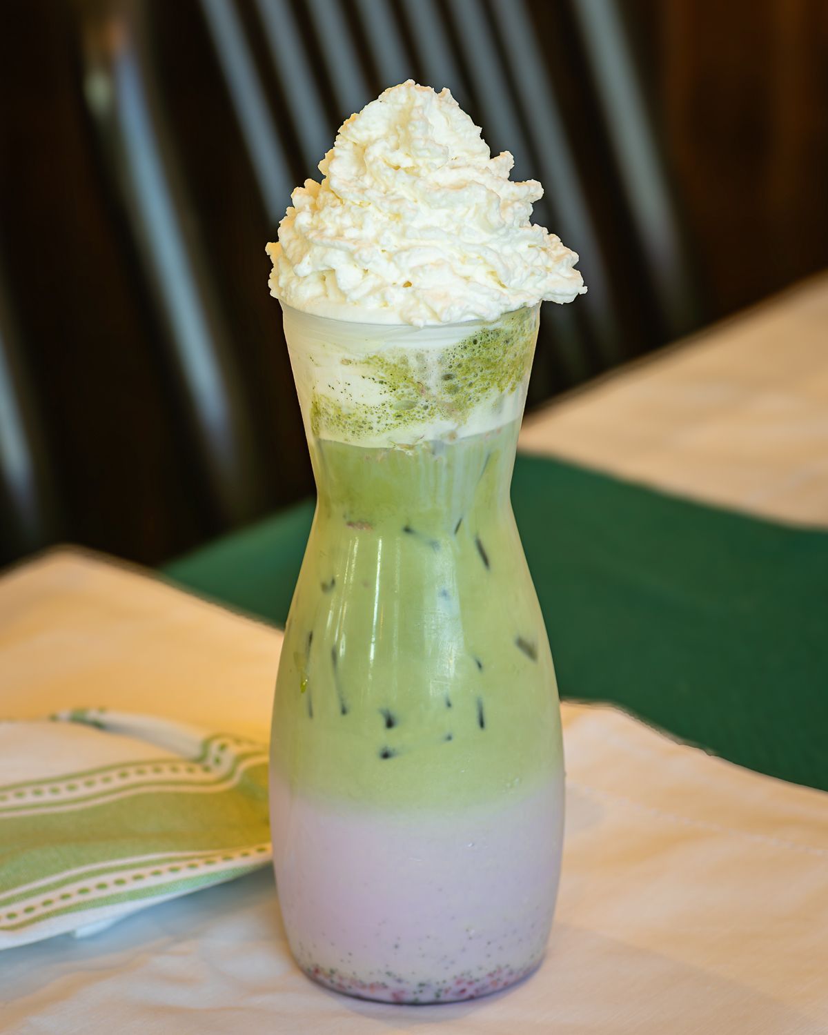 Green matcha drink with purple layer, topped with whipped cream, in tall, hourglass-shaped glass.
