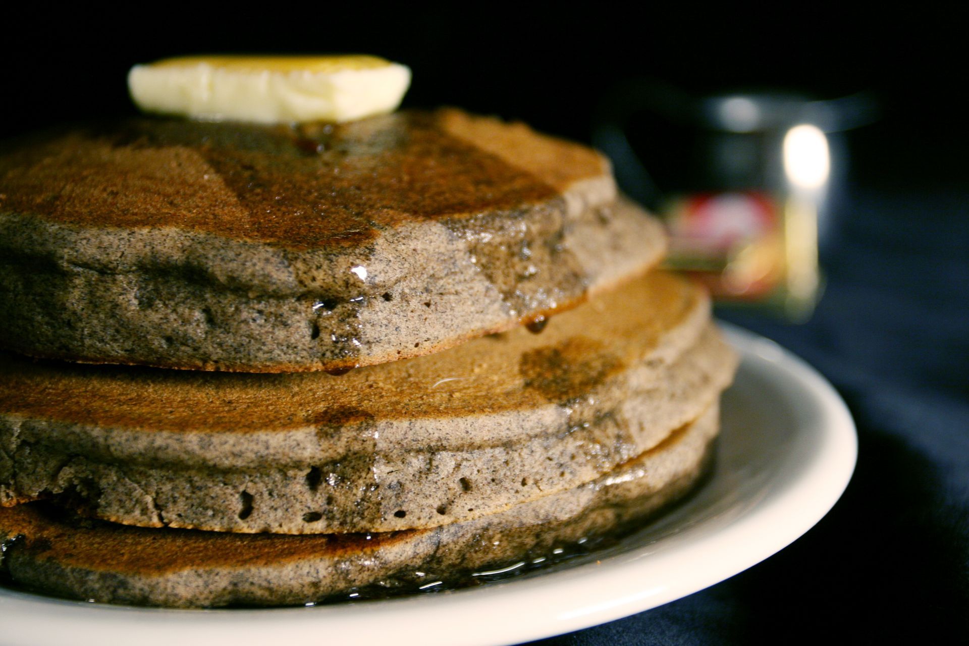 Stack of three dark-colored pancakes drizzled with syrup, topped with butter on a white plate.
