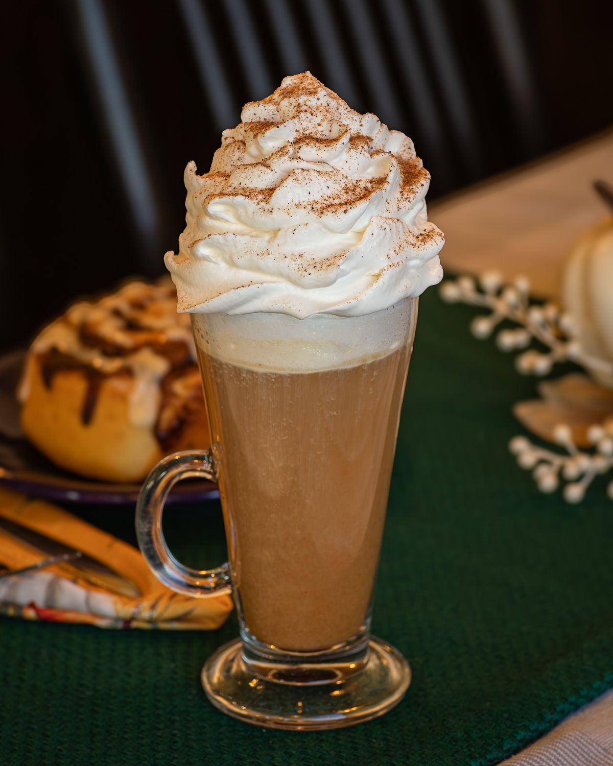 Latte with whipped cream and cinnamon, served in a glass mug; cinnamon roll in the background.