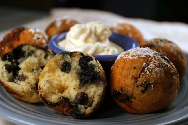 A plate of round, blueberry-filled muffins with powdered sugar and a side of whipped cream in a small blue bowl.
