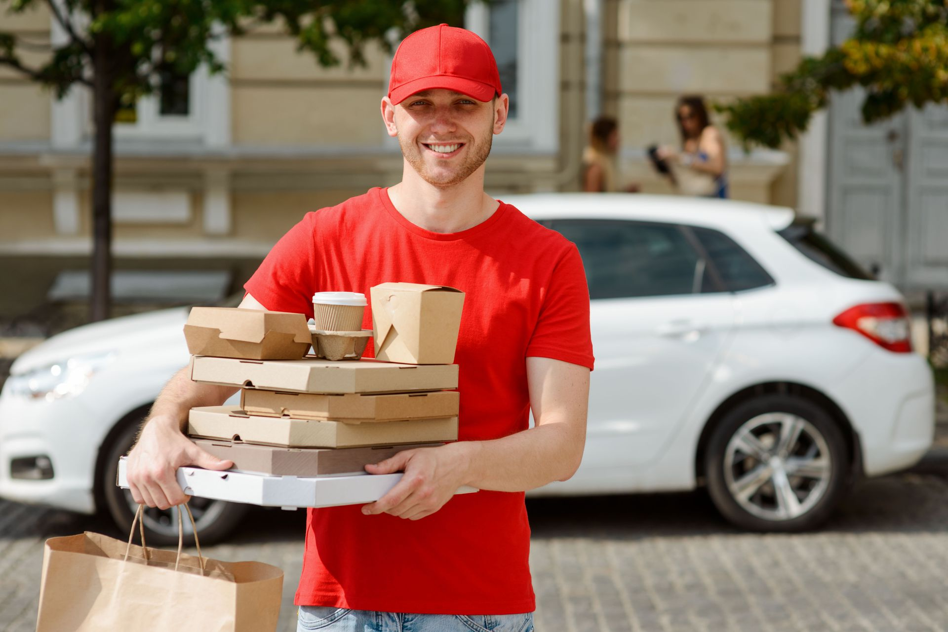 A delivery worker in a red uniform smiles while holding a stack of food containers in front of a white car.