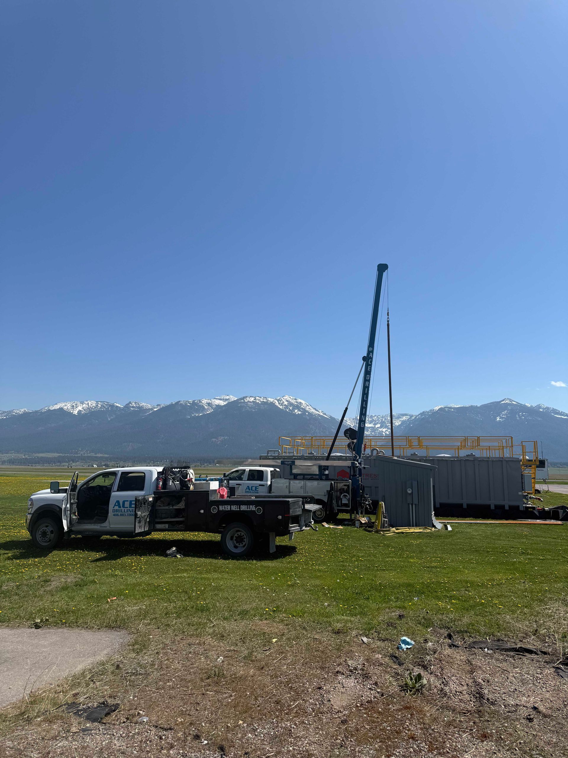 A drilling rig next to a truck, set against a backdrop of snow-capped mountains under a blue sky.