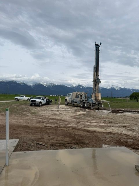 Drilling rig operating in a muddy field, with mountains in the background under a cloudy sky.