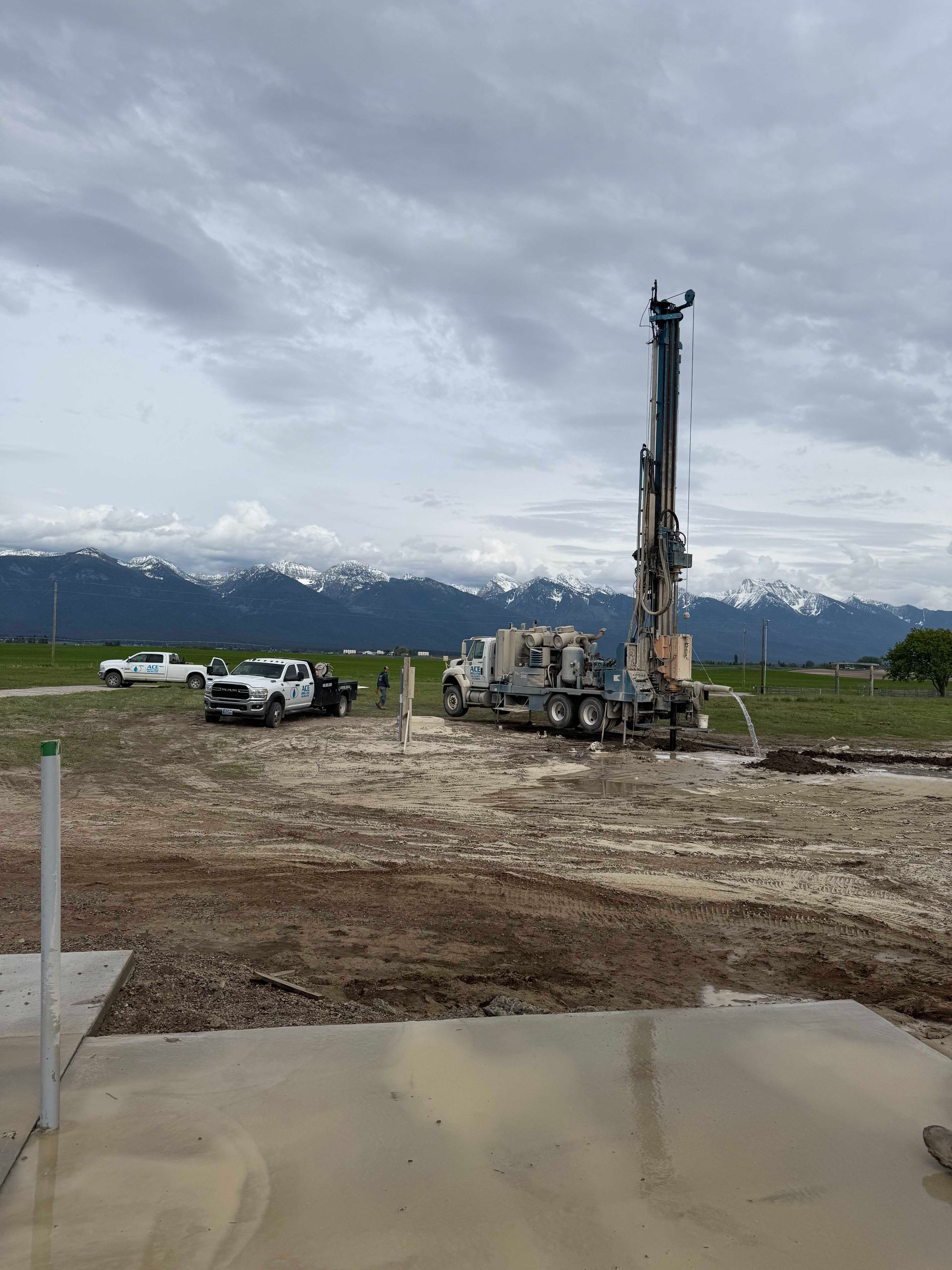 Drilling rig in muddy field with trucks, mountains in background, cloudy sky.