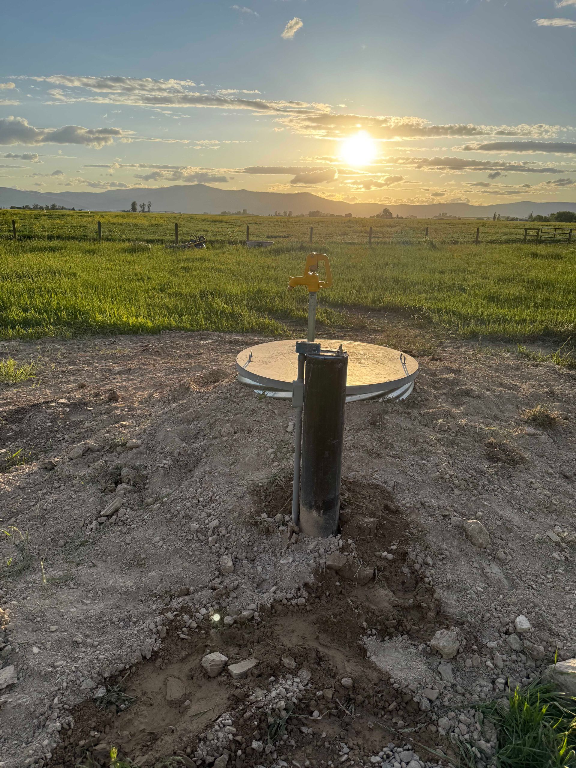 Well pipe in a field at sunset. Green grass, dirt, yellow sun, cloudy sky.