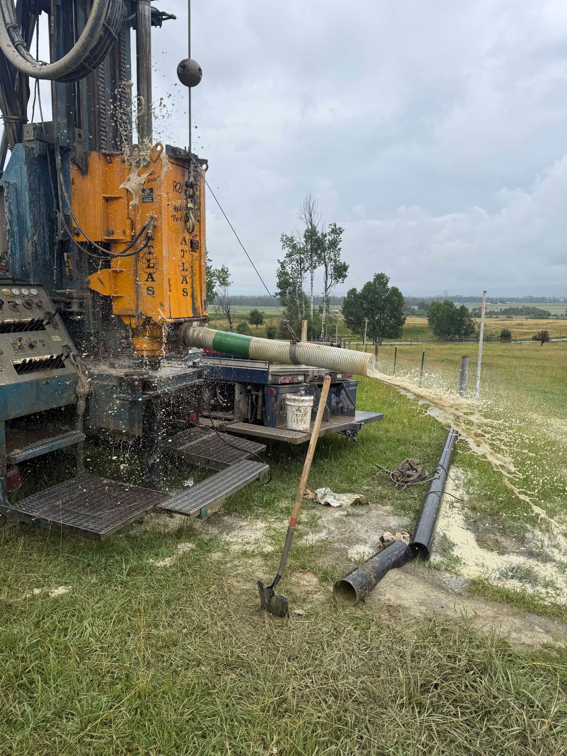 Drilling rig spraying water and debris on a grassy field; cloudy sky in the background.