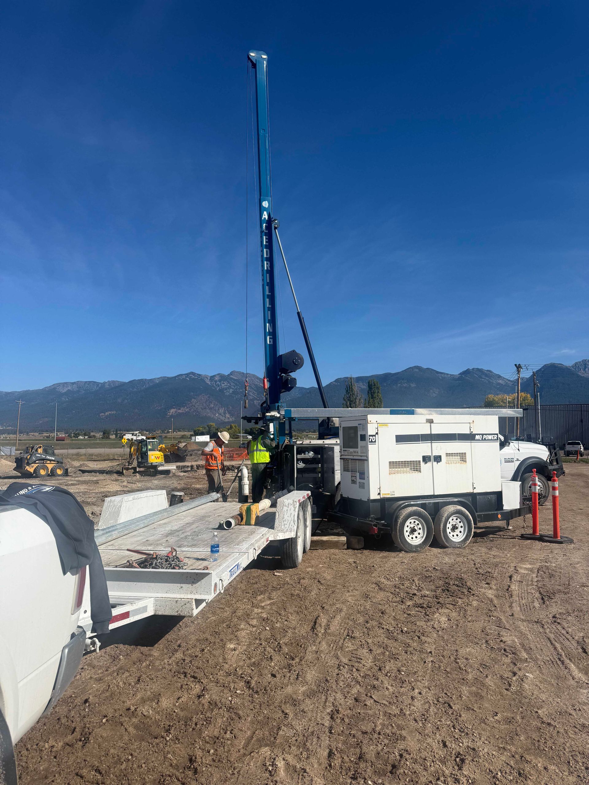 A drilling rig on a trailer in a field with mountains in the background under a blue sky. Two workers are present.