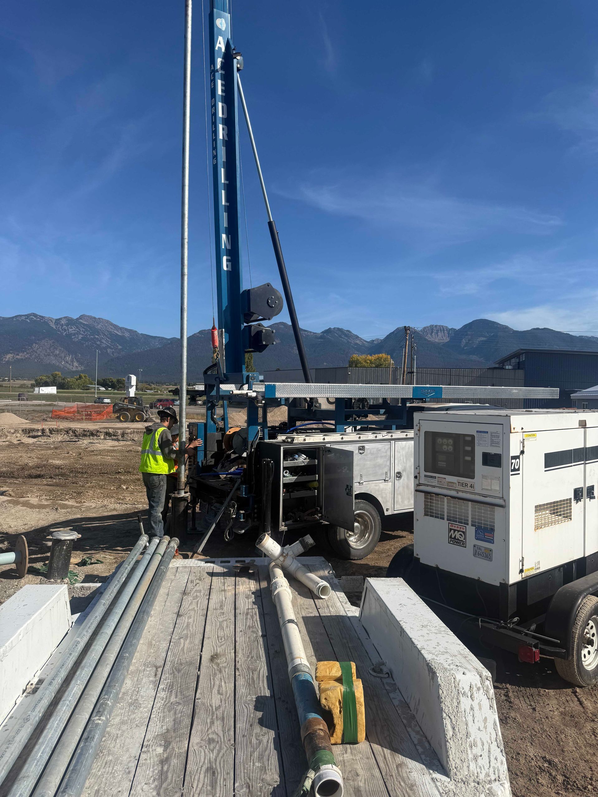 Worker near a blue drilling machine, white generator, and equipment on a flatbed, with mountains in background.