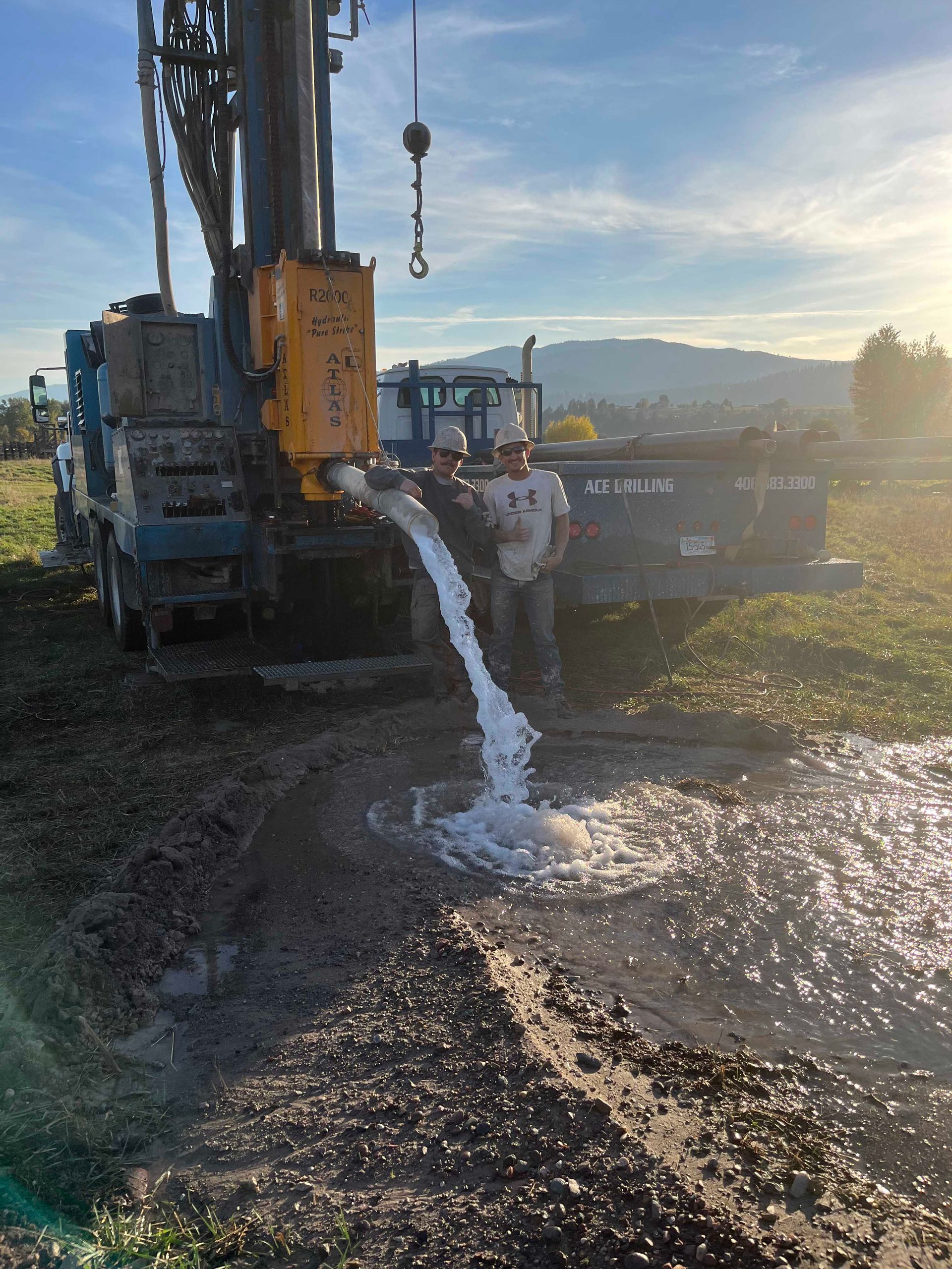 Well drilling rig pumping water onto muddy ground; man stands nearby, mountains in the background.