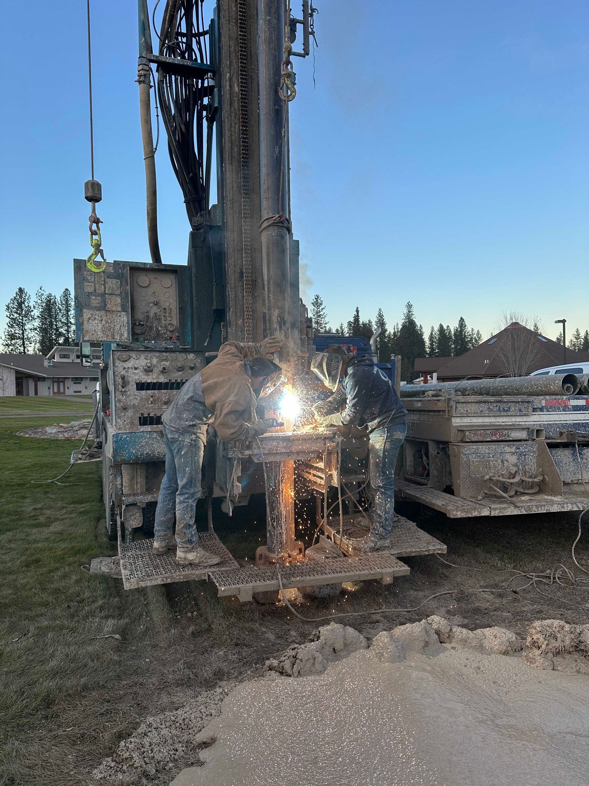 Two people welding on a drilling rig in a muddy field. Sparks and bright light are visible.