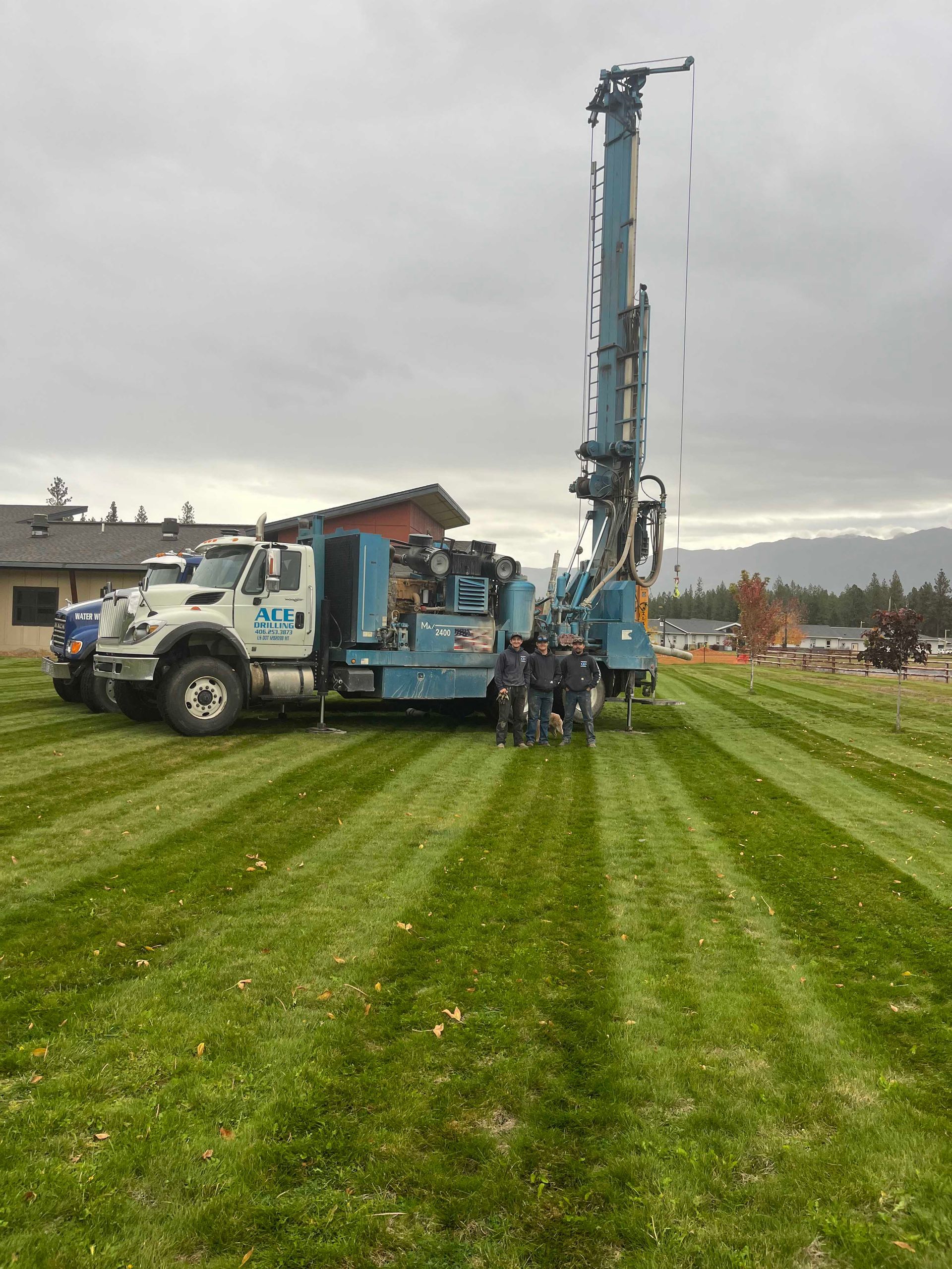 Blue drilling rig with crew on green lawn under overcast sky.