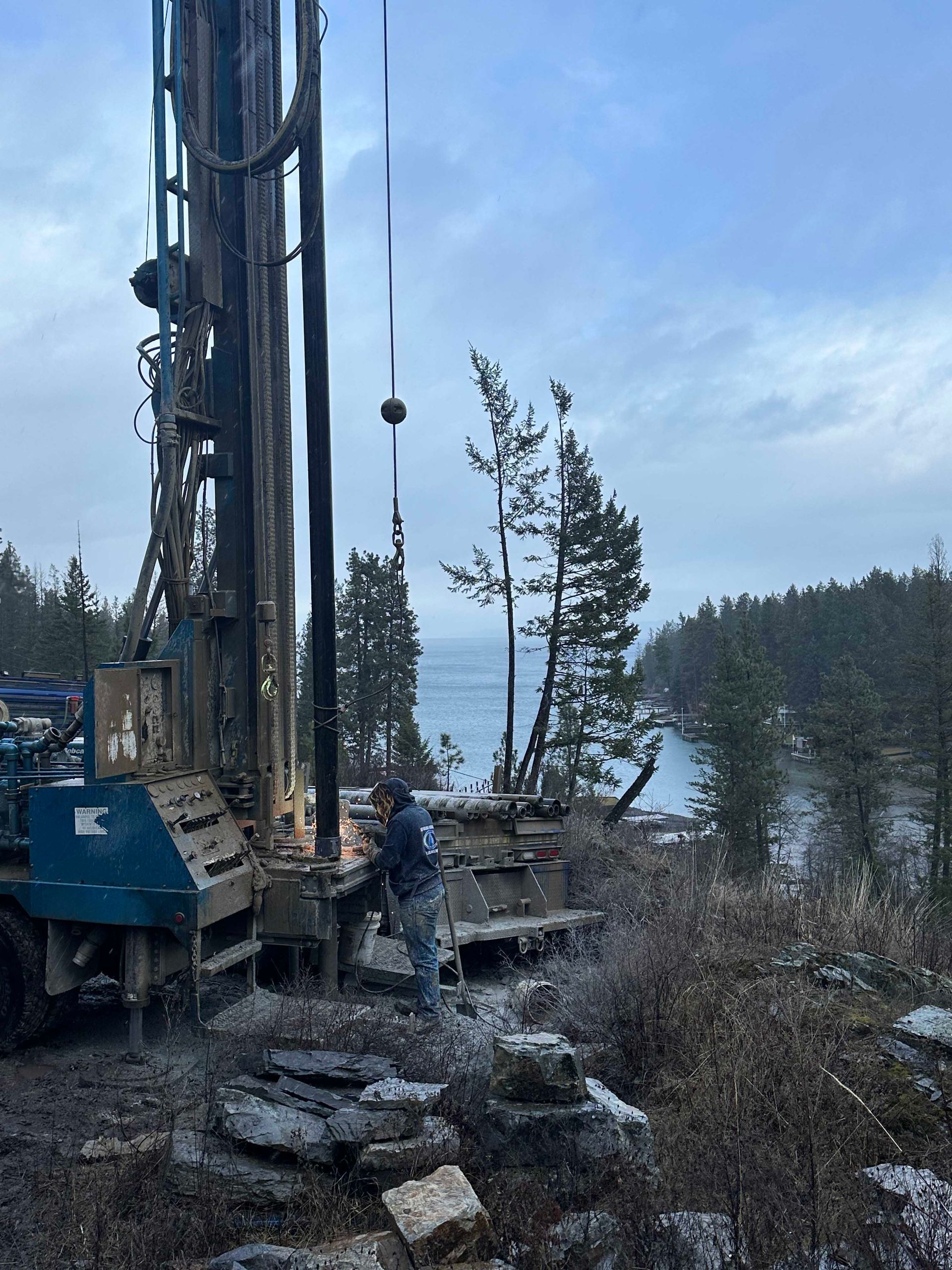 Drilling rig on rocky terrain near ocean. Person works near the equipment under an overcast sky.
