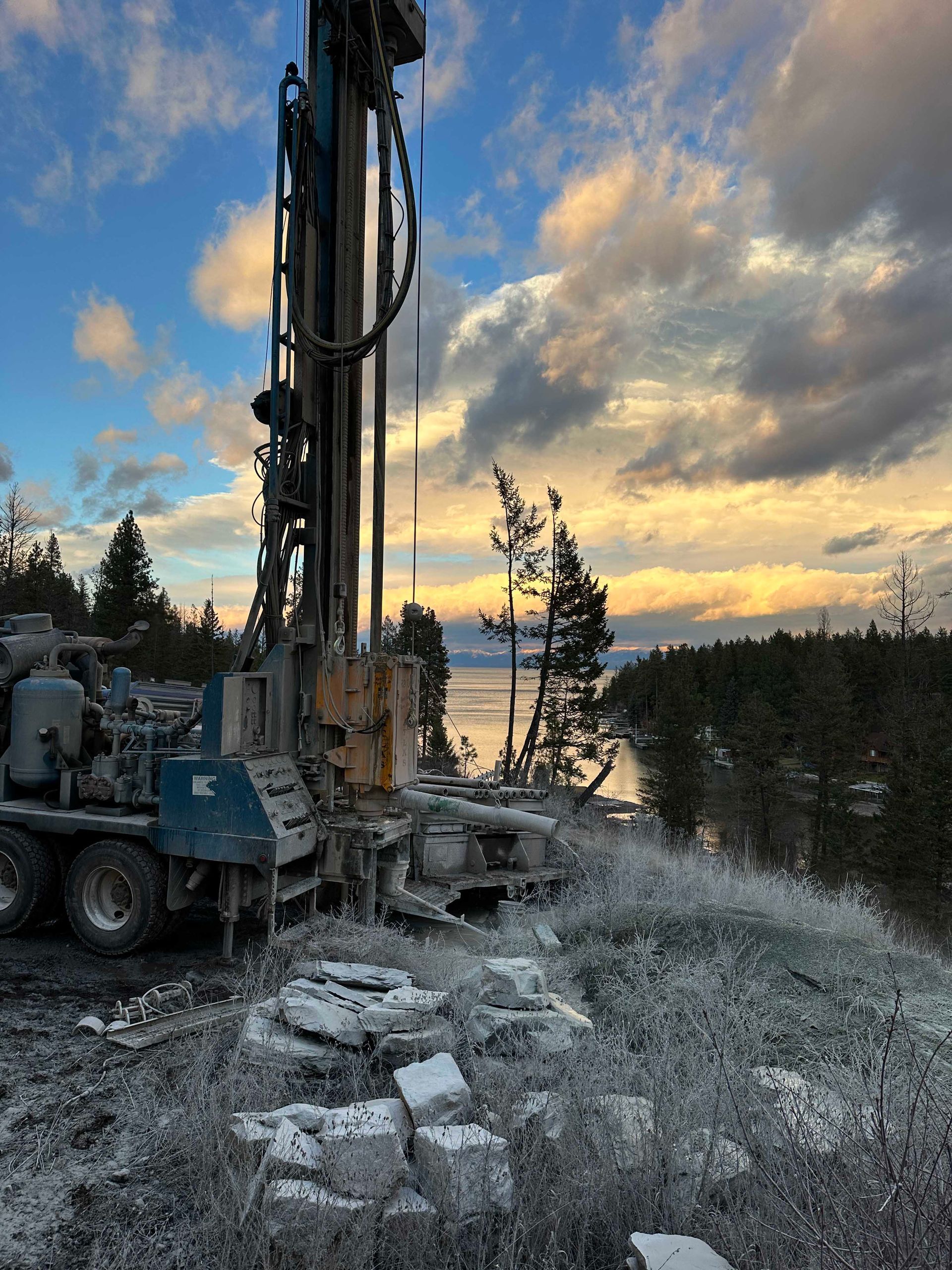 Drilling rig on a hillside overlooking water at sunset. Sky is blue and yellow, ground covered in frost.