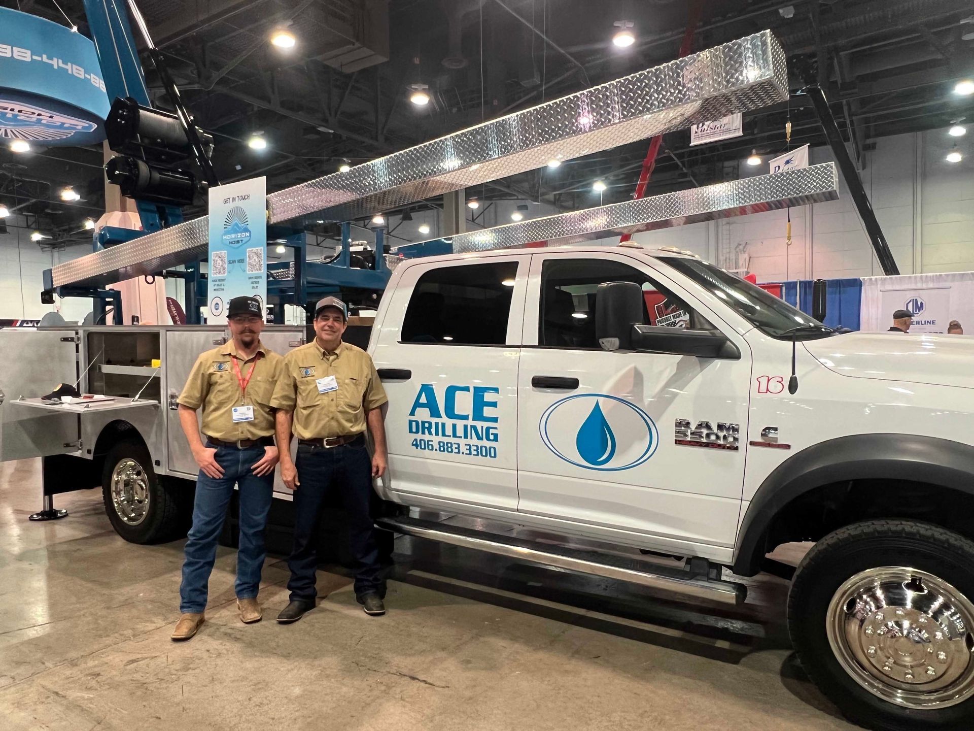 Two men beside a white ACE Drilling truck, a crane in background at a trade show.