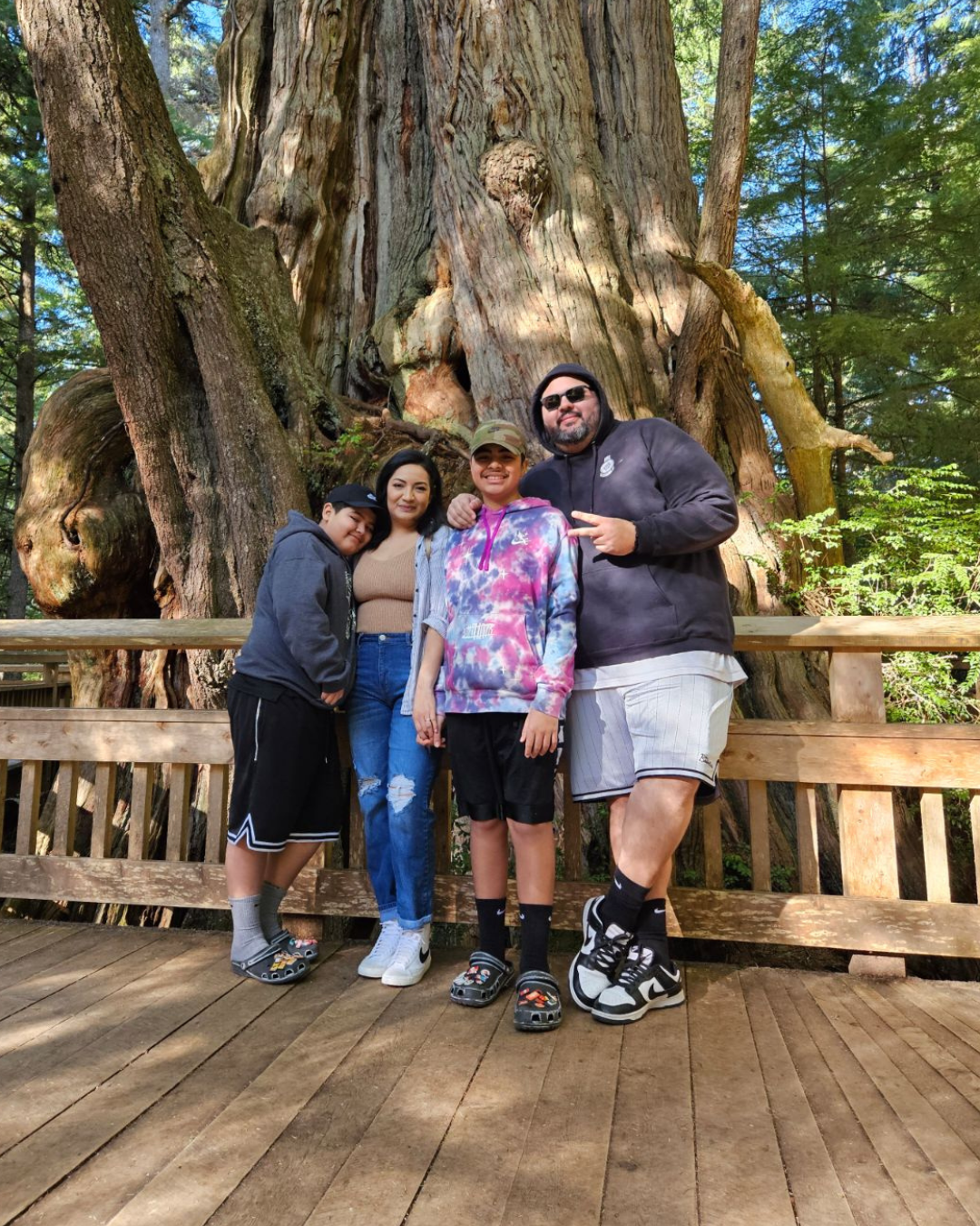 Four people pose on a wooden deck before a giant tree in a sunny forest.