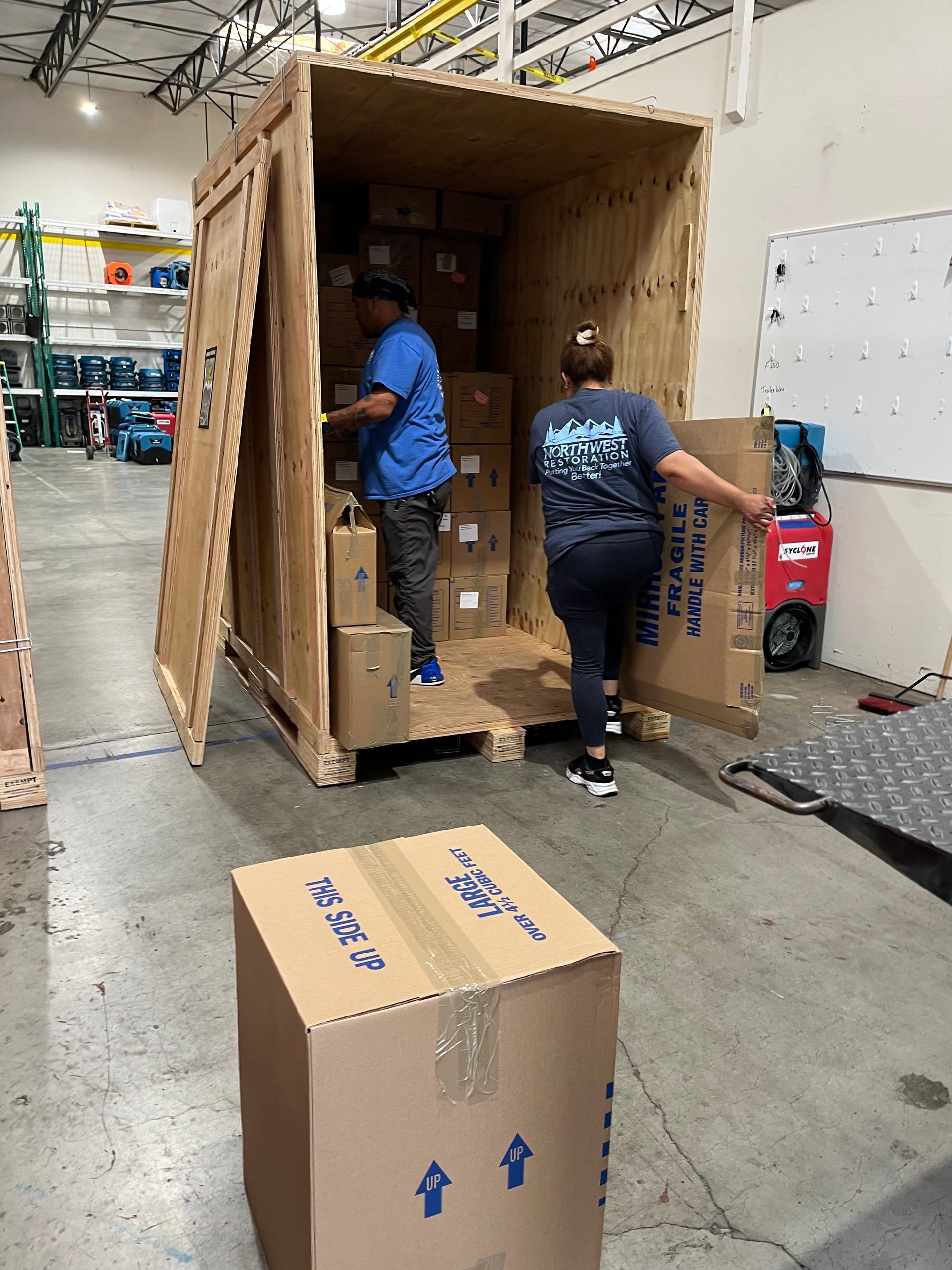 Two workers packing boxes into a large wooden crate in a warehouse.