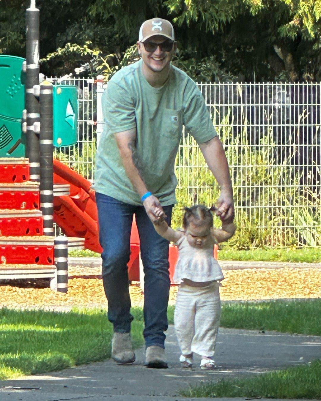 Man in a cap and green shirt helps a toddler walk on a paved path