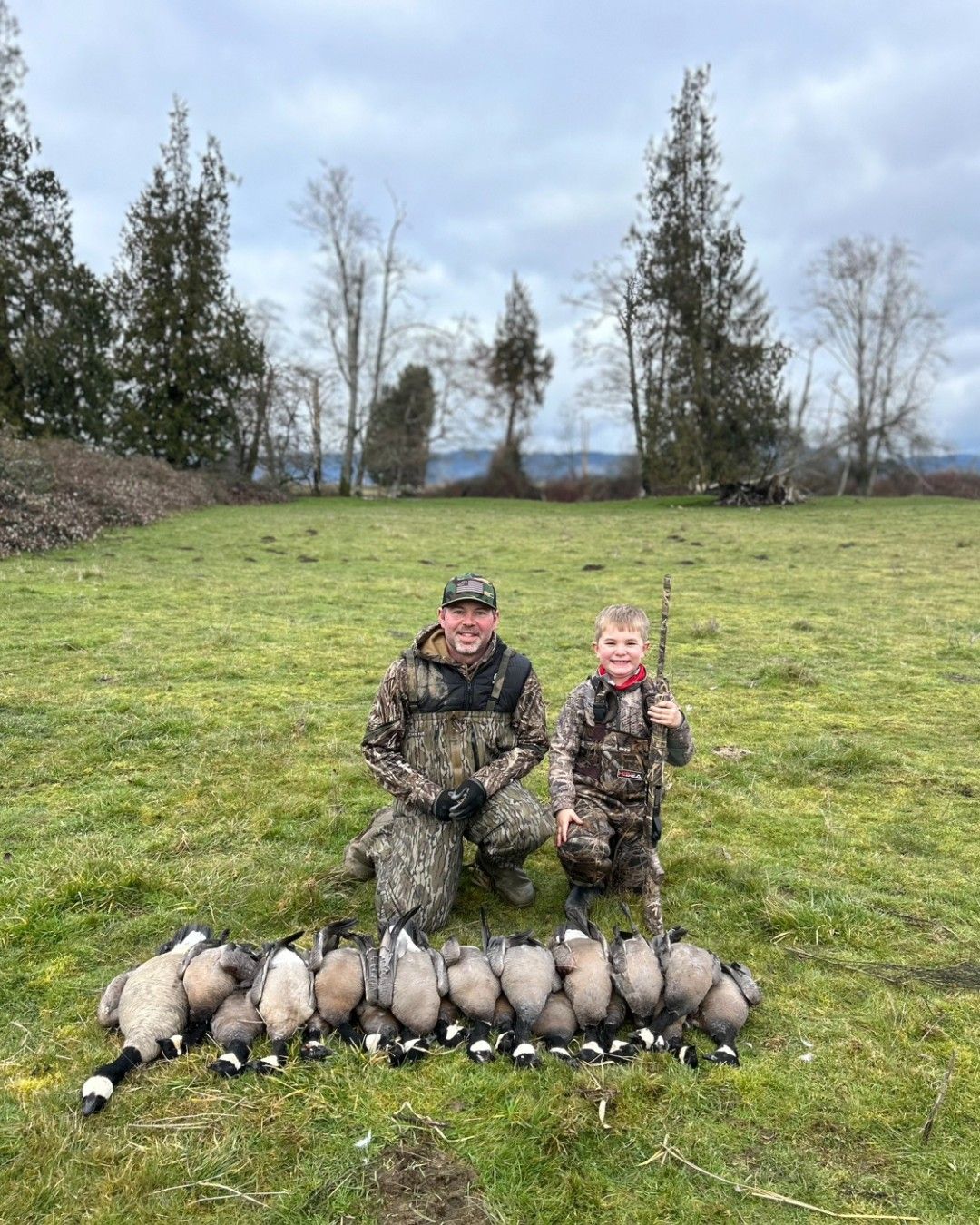 Man and child in camouflage kneel with harvested geese on a grassy field under a cloudy sky.