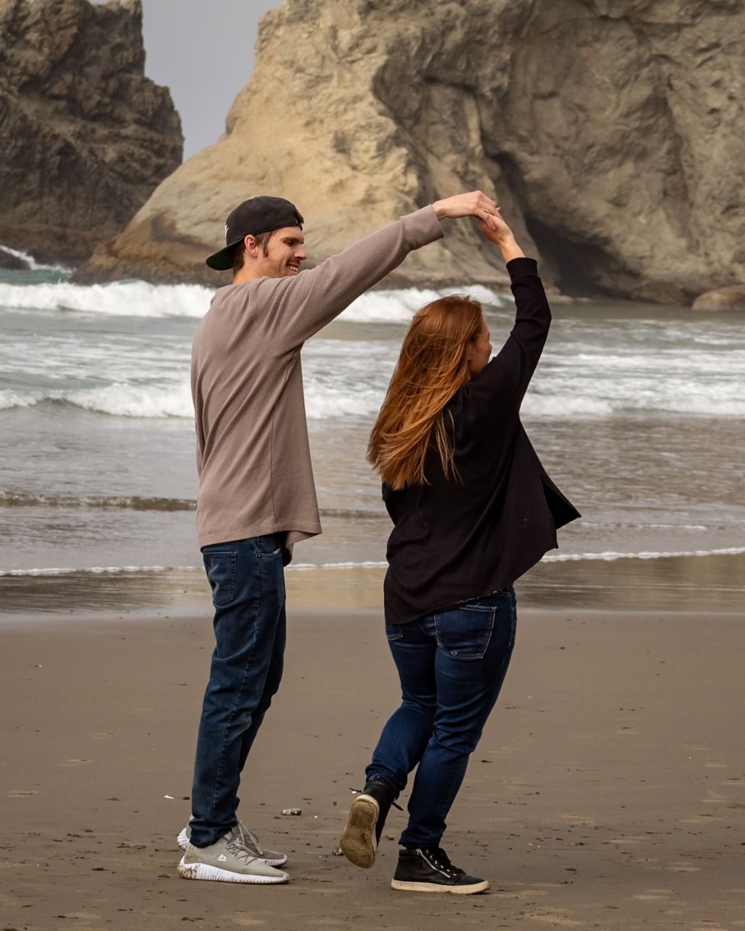 Couple dancing on a beach; holding hands overhead; ocean and rock formations in background.