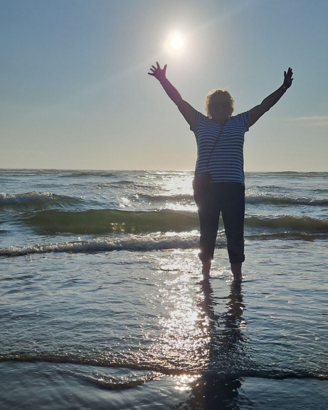 Person standing in ocean with arms raised towards bright sun.