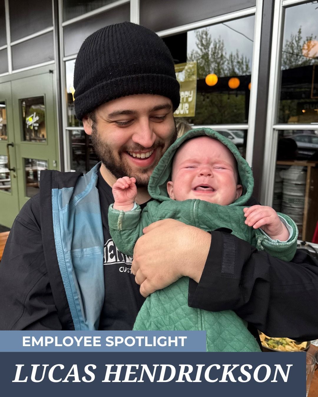 Man in black hat smiles, holding crying baby in green hooded onesie