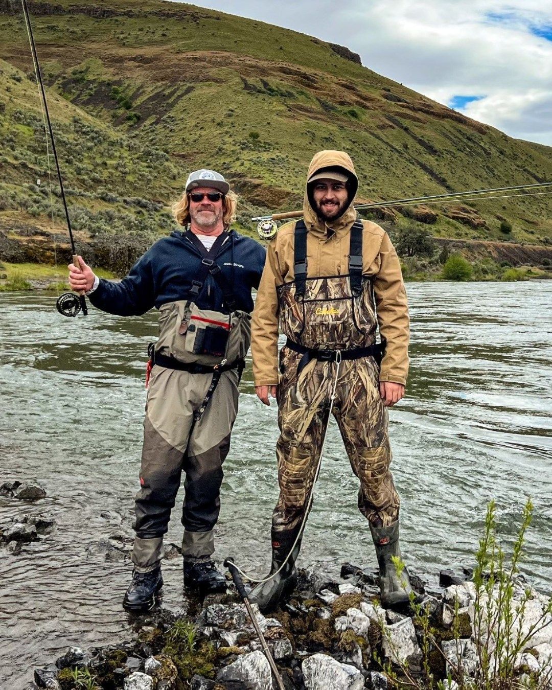 Two people in waders fly fish in a river with a green hillside in the background.