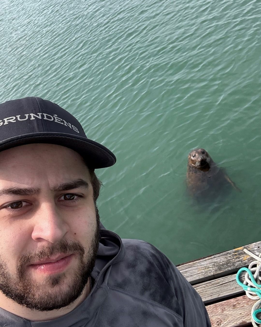 Man taking selfie with a seal in water, on a wooden dock.