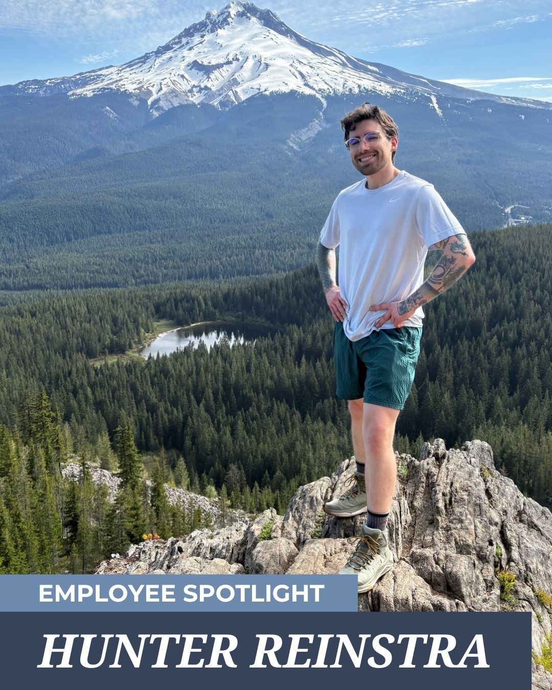 A smiling person stands on a rocky mountain ledge overlooking a pine forest and a snow-capped peak. 