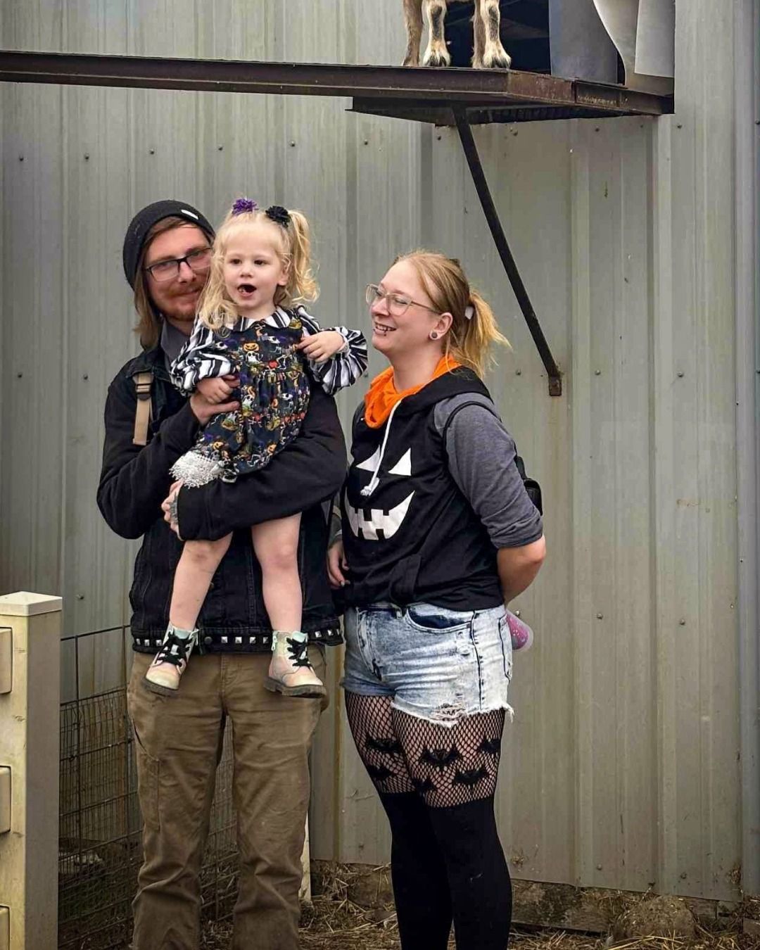 A family stands smiling in front of a metal wall; the child is held, and the parent on the right wears a pumpkin vest. 