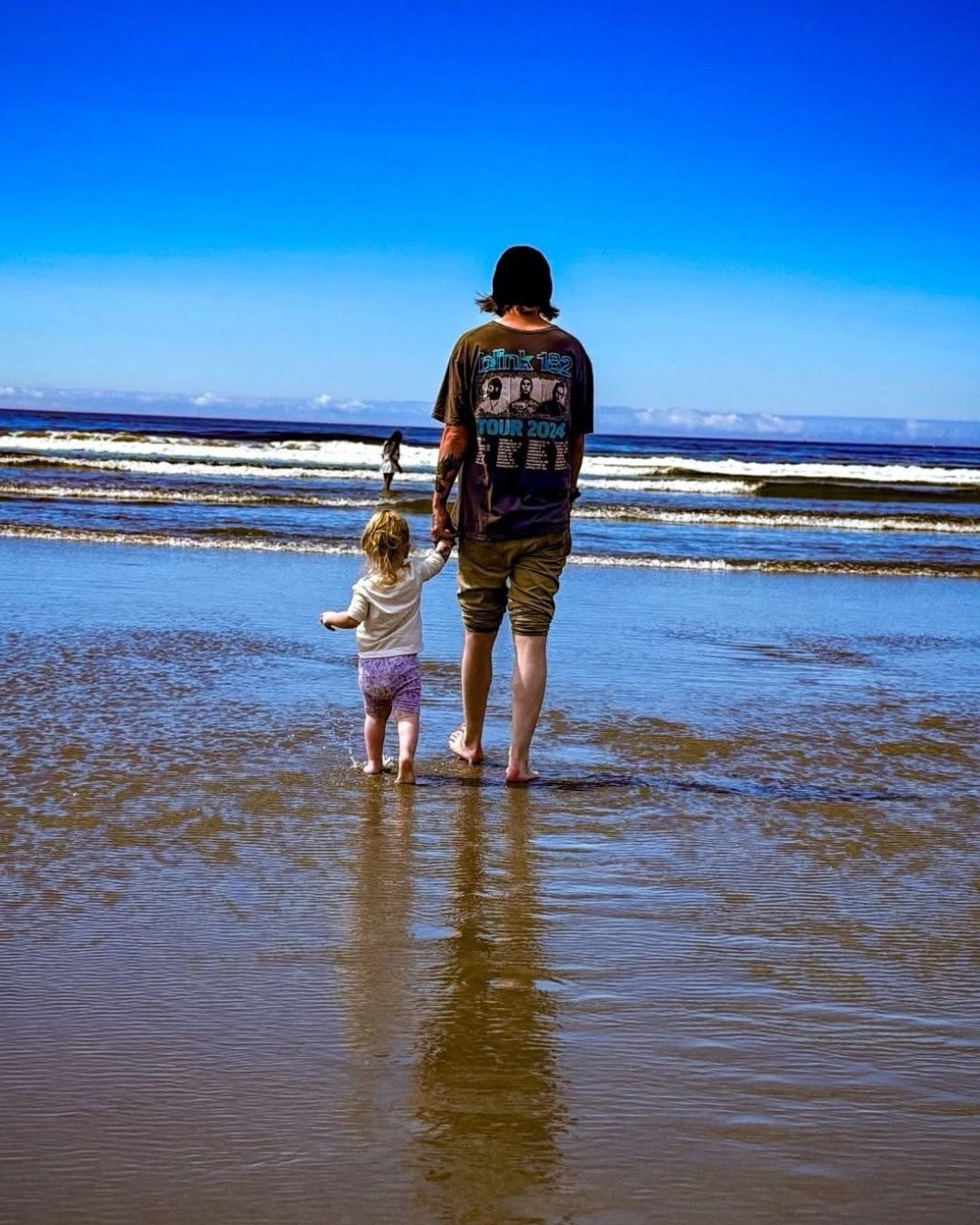 A person and a toddler walk hand-in-hand through shallow ocean water toward the waves under a clear blue sky. 