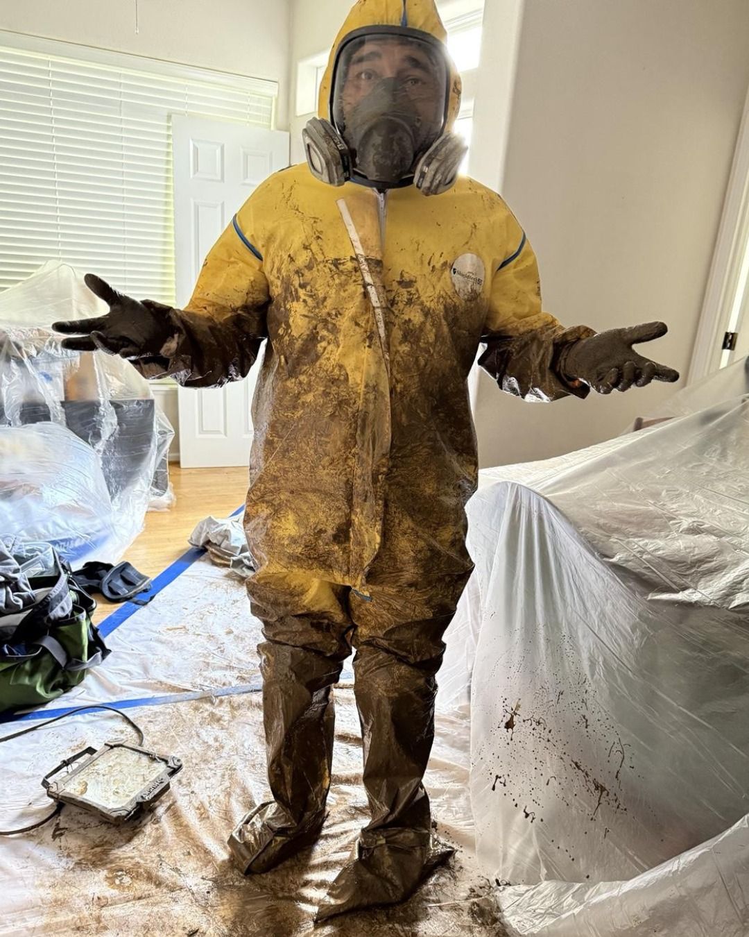 A person in a yellow hooded hazmat suit and respirator covered in mud, standing in a room covered with plastic sheeting. 