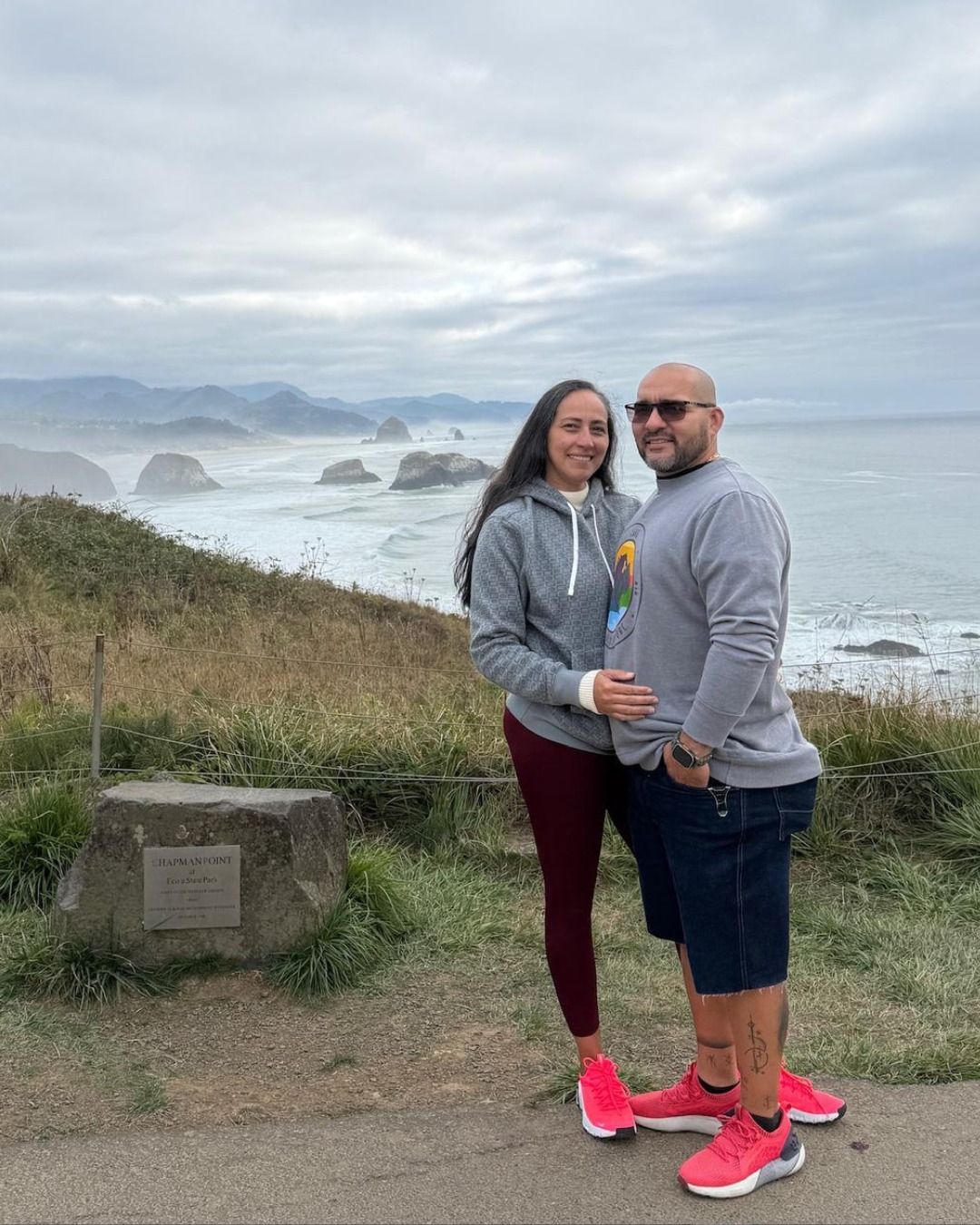 A couple stands side-by-side smiling on a coastal hillside overlooking the ocean and rocky islands under a cloudy sky. 