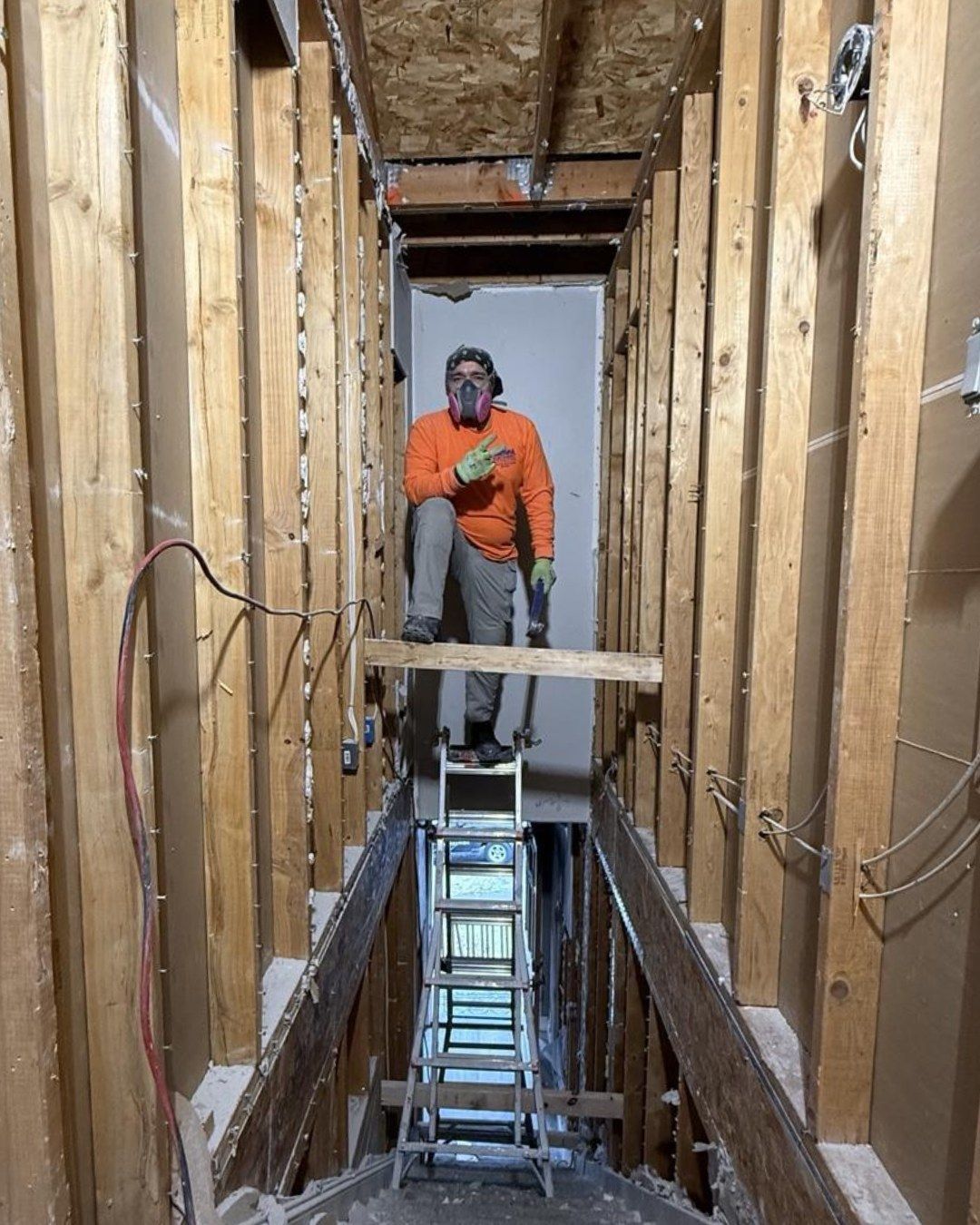 A construction worker in an orange shirt stands on a wooden plank balanced on a ladder between wall studs in a narrow space. 
