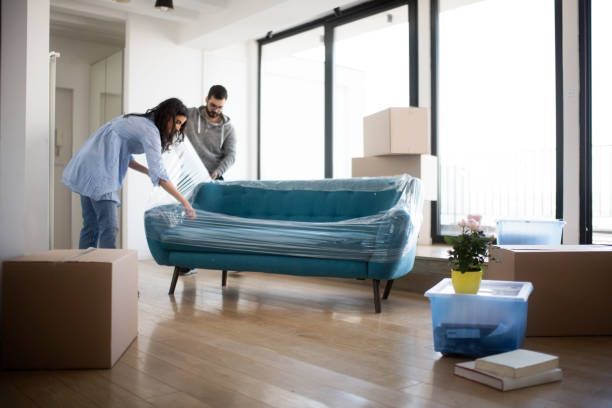 Couple wrapping a teal sofa in plastic in a new home, surrounded by moving boxes.