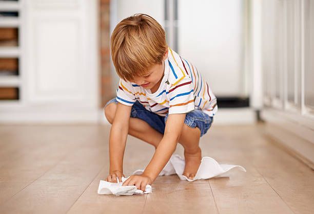 Child cleaning a wooden floor with a white wipe, smiling.