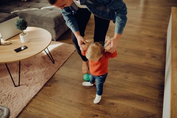 A person assists a toddler in taking steps on a wooden floor in a living room.