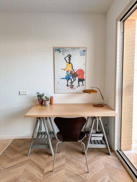 Desk with painting and chair near window. Wooden desk on metal sawhorse legs, artwork depicts figures.