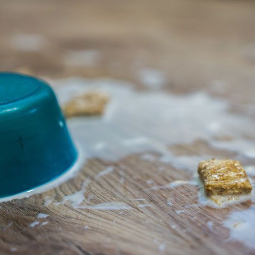 Overturned blue bowl on a wooden surface with spilled white liquid and cereal pieces.