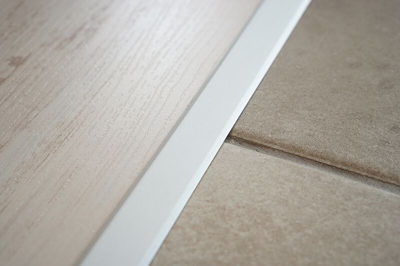 White molding strip separating light wood flooring from beige tile.