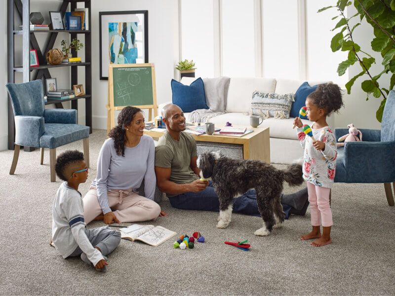 Family playing with a dog in a living room, smiling. A child holds a toy.
