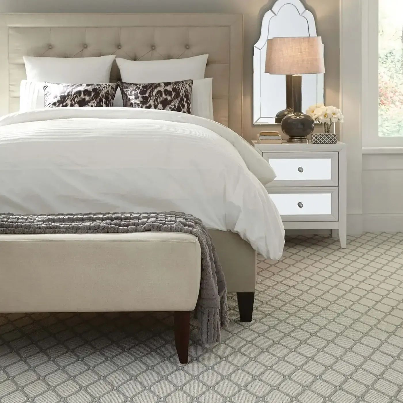 Bedroom with tufted headboard, white bedding, patterned carpet, bench at foot of bed, and nightstand with lamp.