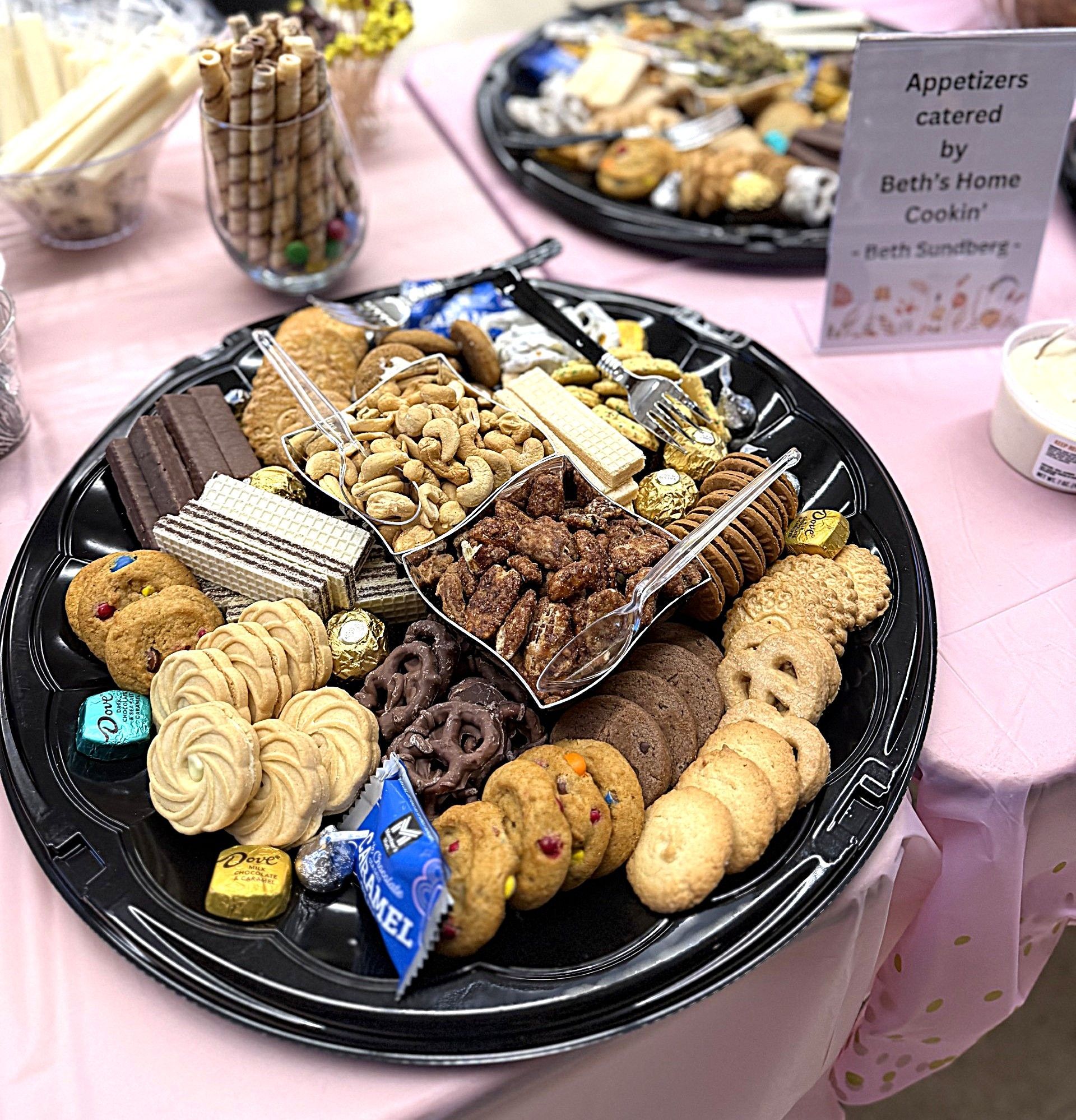 Assorted cookies and treats on a black platter at a dessert table with a pink tablecloth.