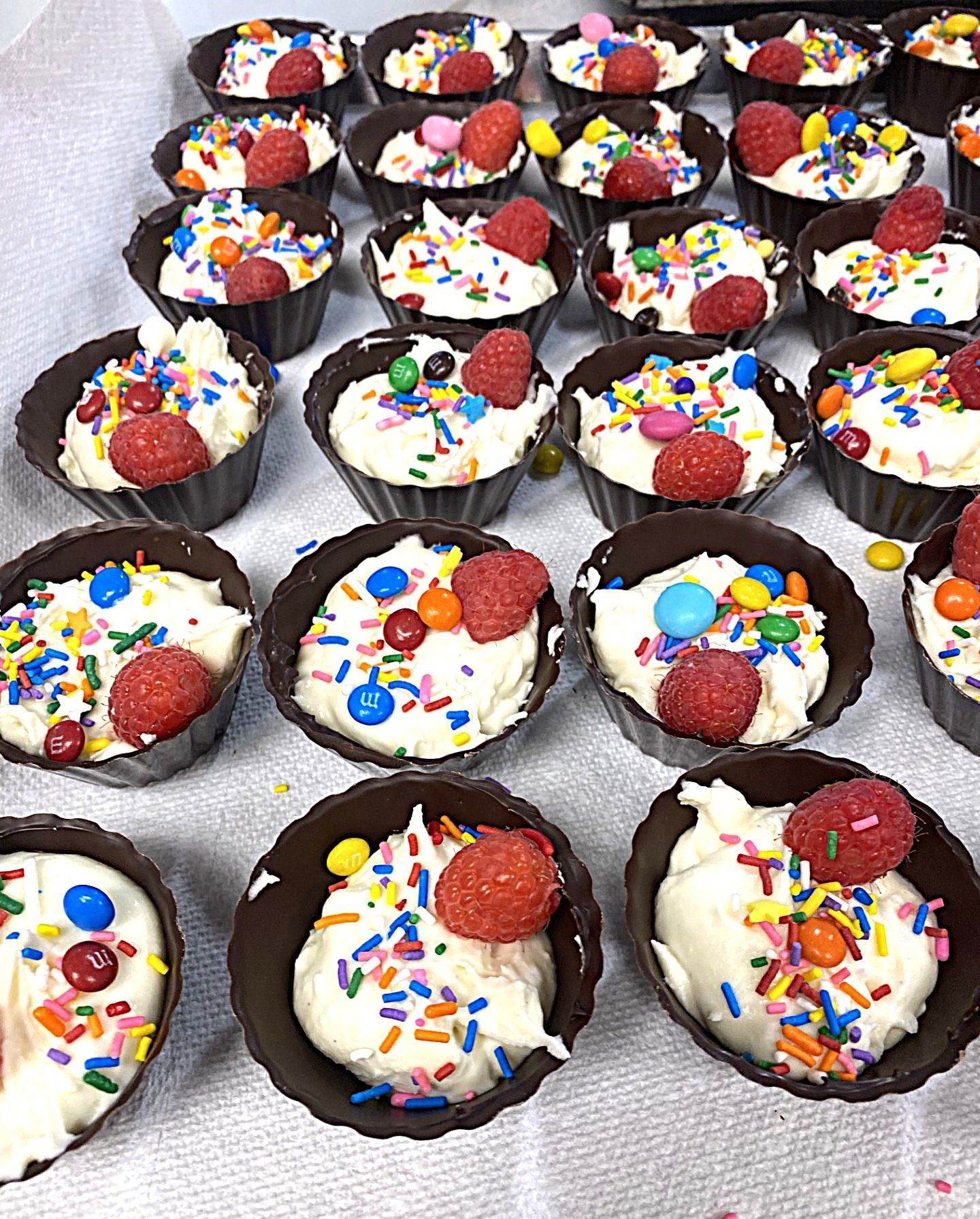 Chocolate dessert cups topped with white cream, strawberries, and colorful sprinkles on a tray