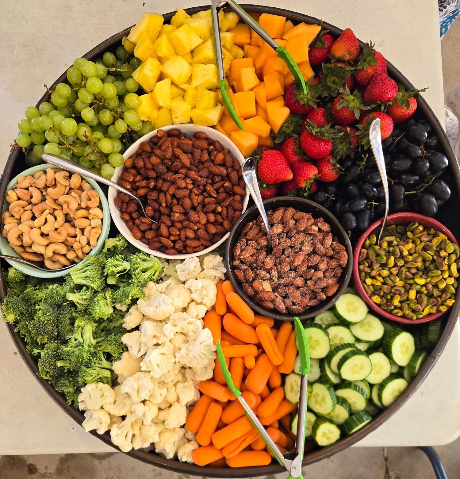 Assorted fruits, nuts, and vegetables arranged in a circular tray, including grapes, mango, strawberries, berries, cucumbers, and carrots.