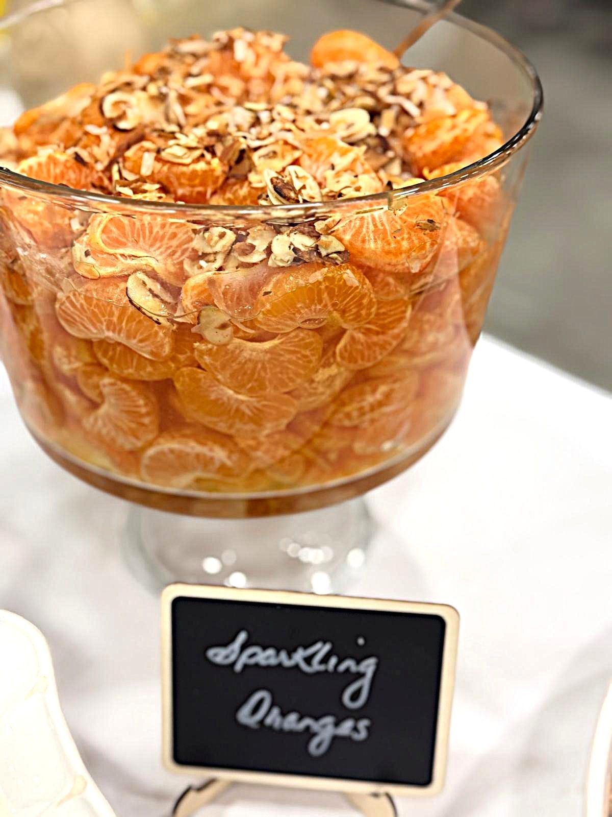Glass dessert cups of orange sparkling grapes topped with sliced almonds and cocoa powder, labeled on a stand.
